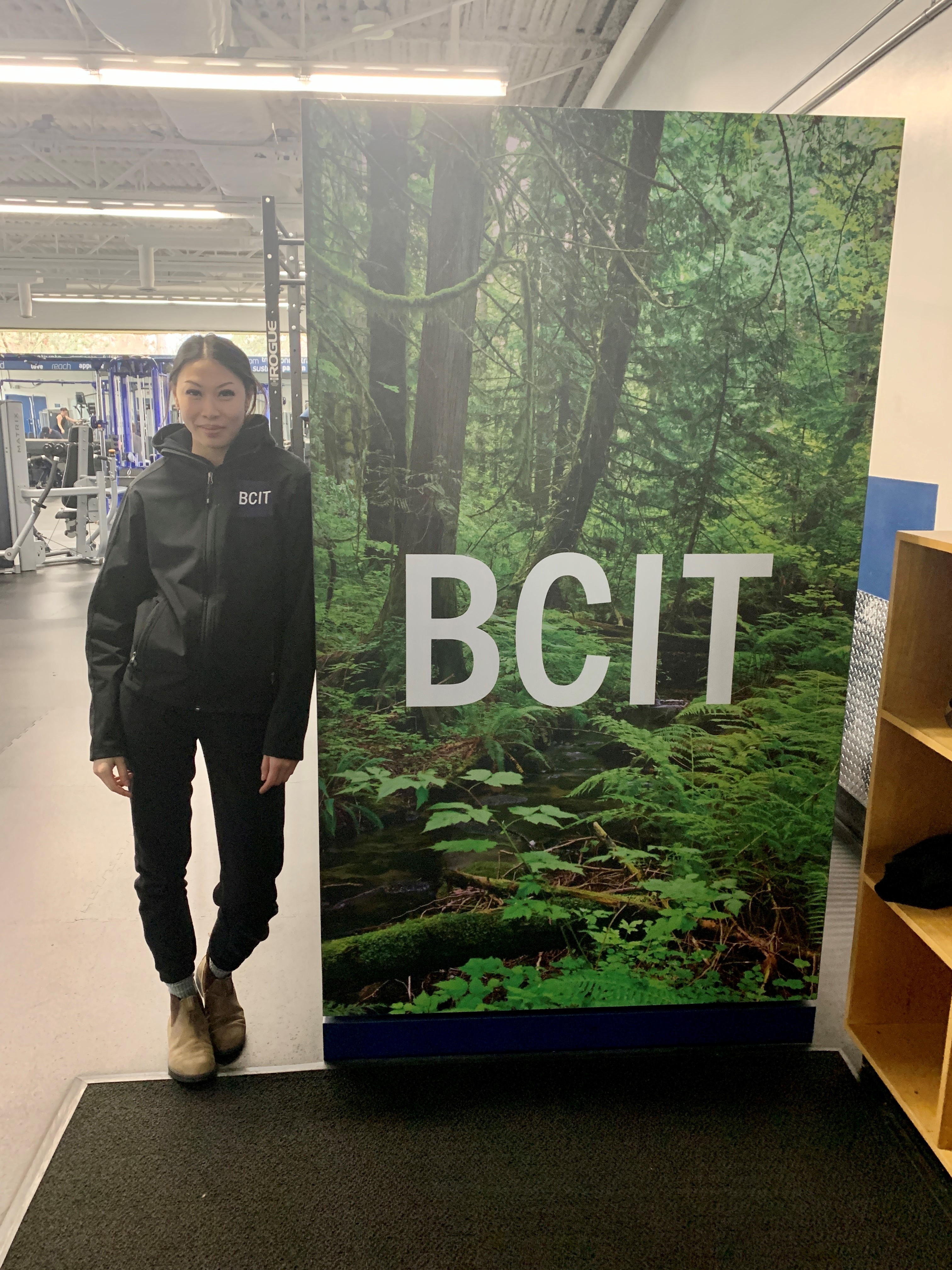 Woman posing beside BCIT poster with gym equipment in the background